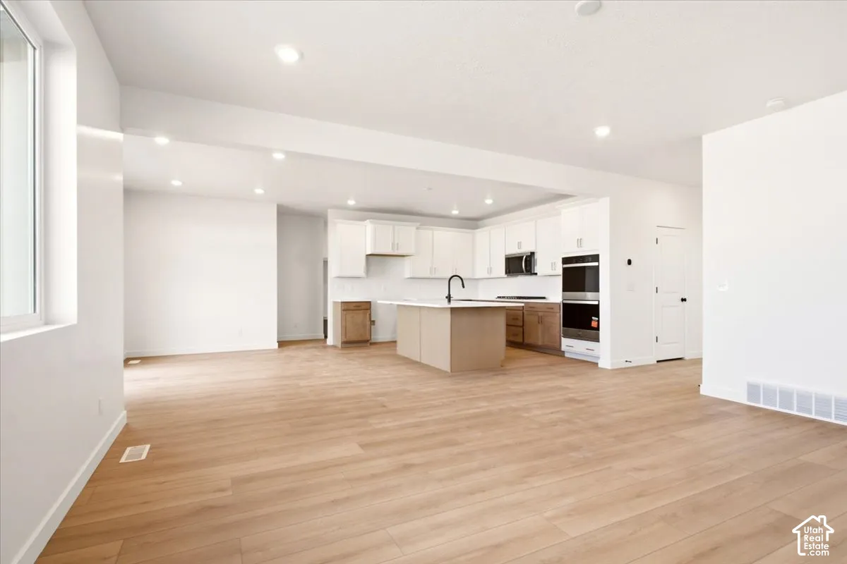 Open-concept kitchen featuring wood-finish flooring, a central island with a matte black faucet, white shaker-style cabinetry, and built-in stainless steel appliances
