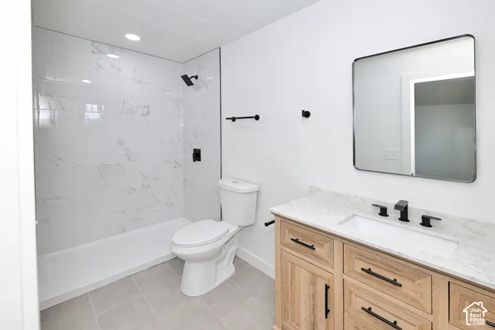 Bathroom featuring stone-pattern tile shower surround, light wood-finish vanity with stone countertop, rectangular undermount sink, matte black hardware, and framed wall mirror