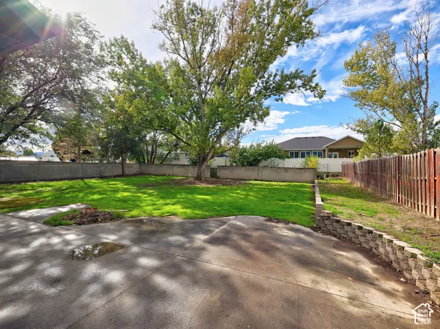 Backyard featuring a manicured lawn, concrete patio, segmented block retaining wall, perimeter wood fencing, and mature trees