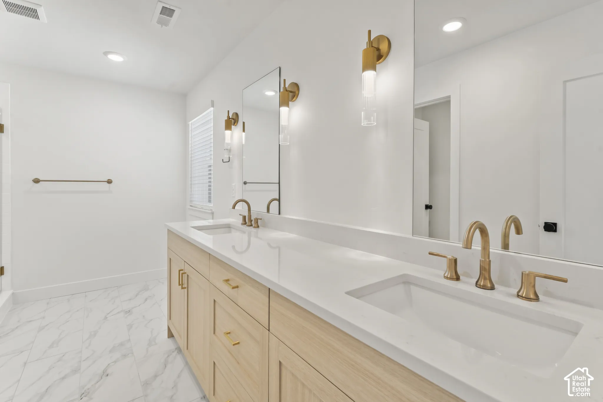 Dual-vanity cabinet with light wood finish, white stone countertop, undermount sinks, brass-toned fixtures, and wall-mounted cylindrical sconces