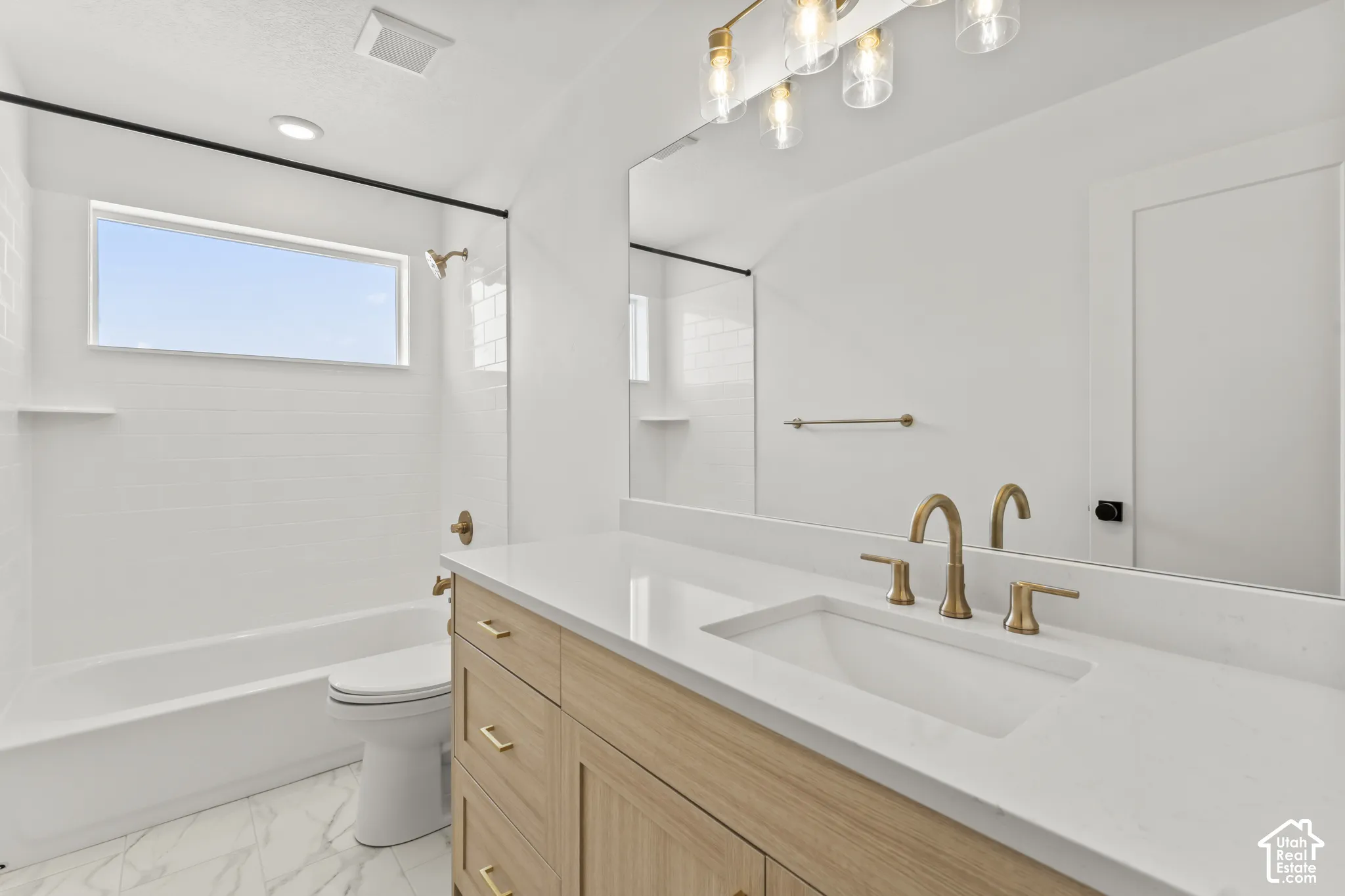 Bathroom featuring a vanity with light wood-finish cabinetry, a white countertop, an undermount sink with brass fixtures, a full-width mirror, and a tub-shower combination with a glass panel