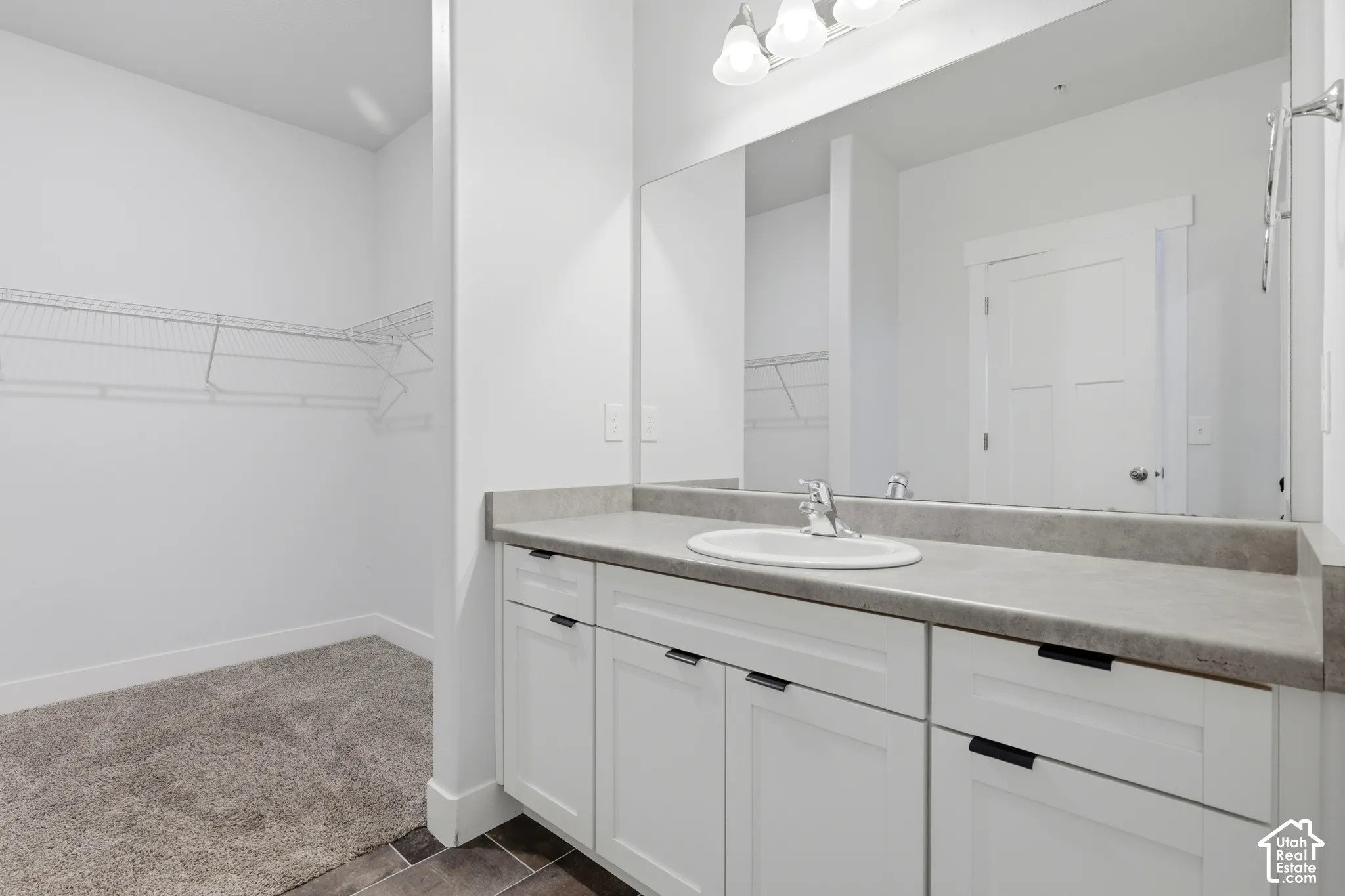 Bathroom vanity featuring white cabinetry, a neutral countertop with an integrated oval sink, a full-width vanity mirror, a three-bulb light fixture, and an open walk-in storage area