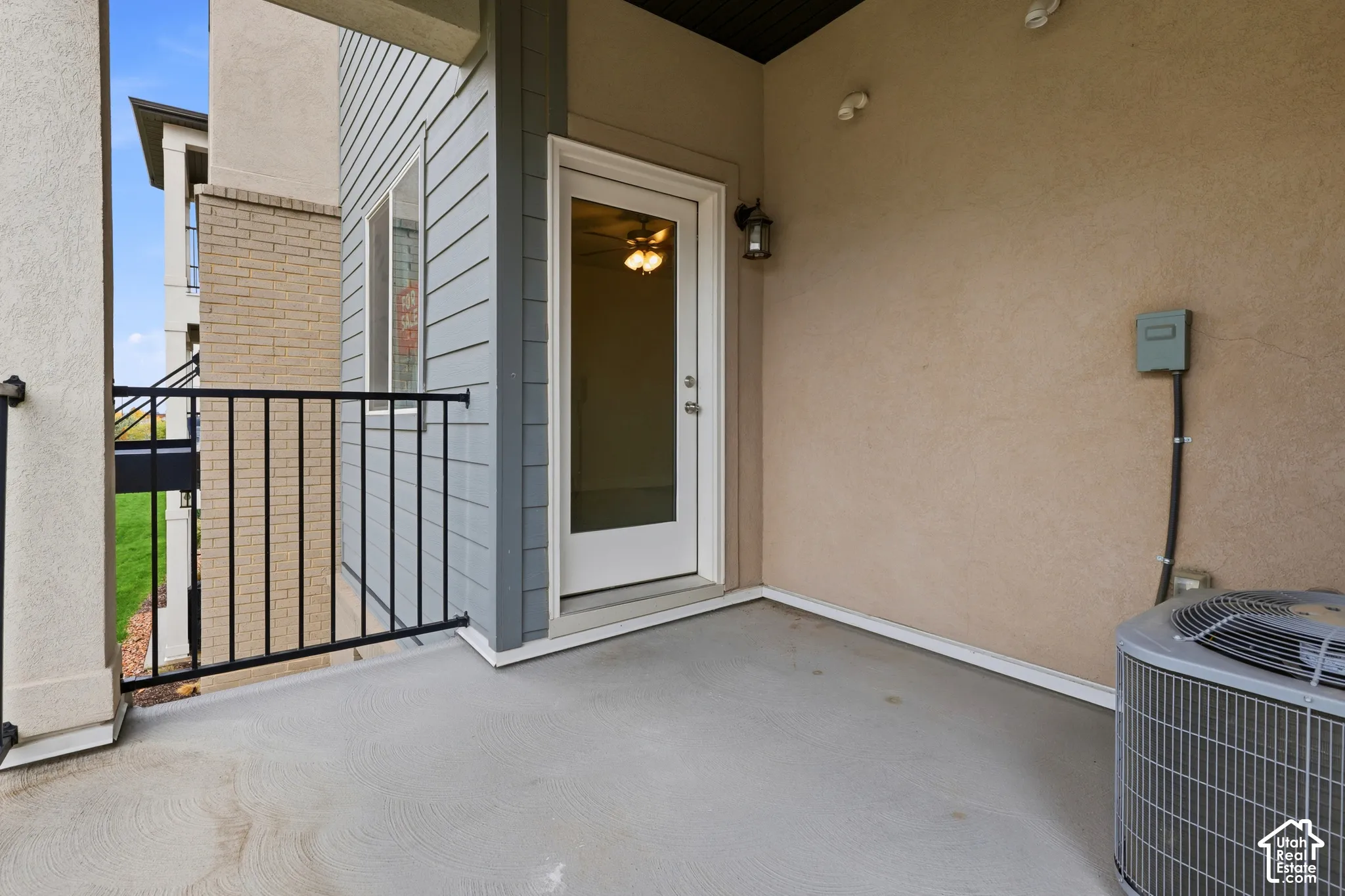 Covered balcony featuring concrete flooring, black metal railing, neutral stucco wall finish, and an exterior wall-mounted light fixture
