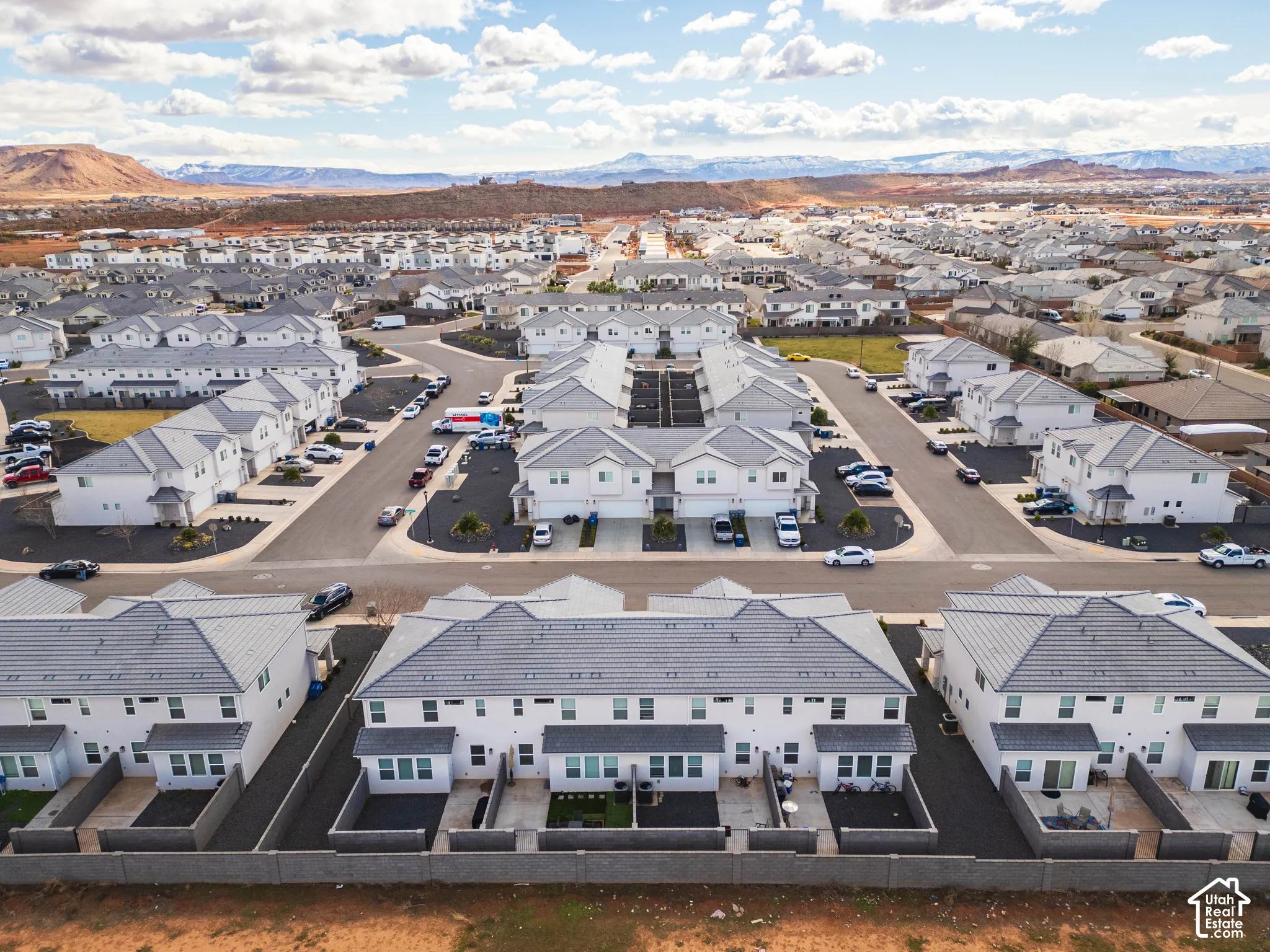 Multi-unit residential complex featuring uniform light-toned exteriors, shingled roofing, paved streets, and private rear patios enclosed by masonry walls with mountain backdrop