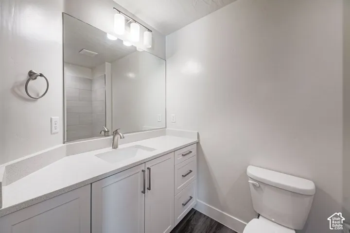 Bathroom featuring a white vanity cabinet with quartz countertop, undermount sink, expansive wall-to-wall mirror, multi-bulb vanity lighting, and wood-finish flooring