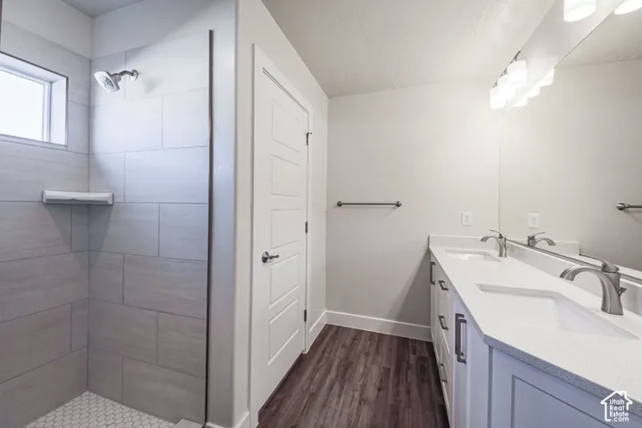 Bathroom featuring a dual-sink vanity with a solid-surface countertop, wood-finish flooring, a full-width mirror, and a walk-in shower with large-format tile surround