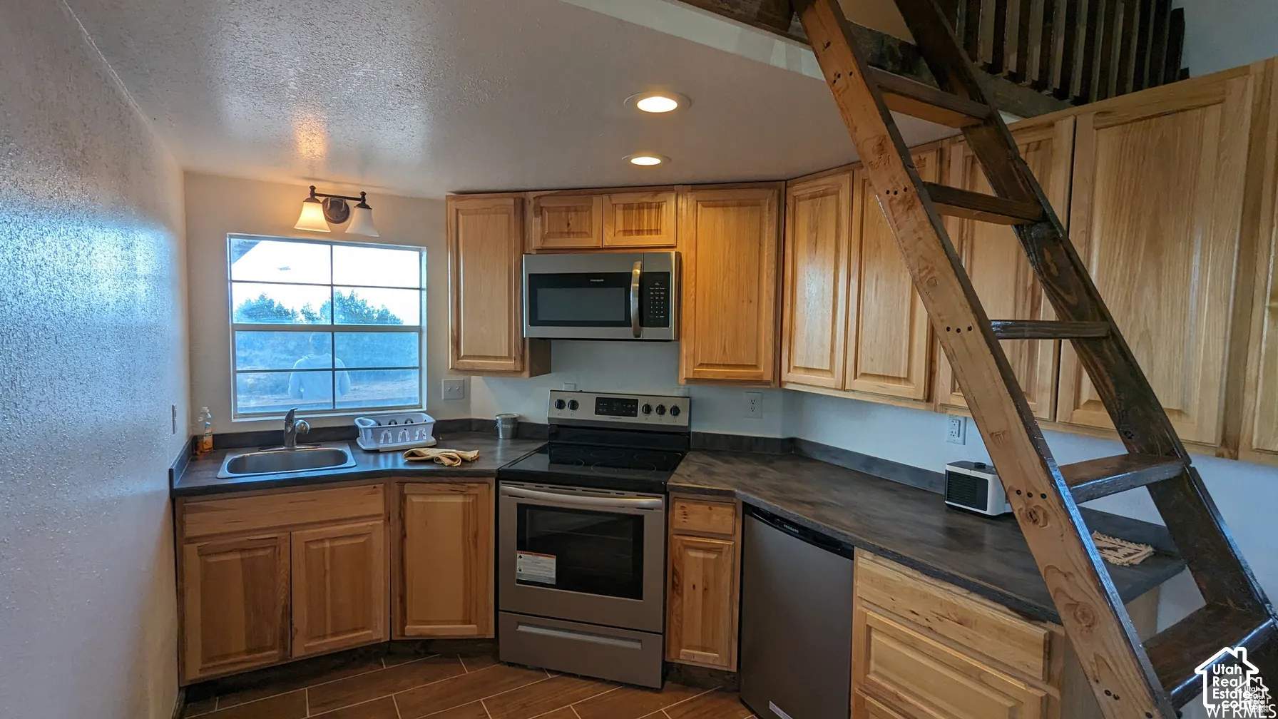 Kitchen featuring light wood cabinetry, dark laminate countertops, stainless steel appliances, tile flooring, and an integrated wooden ladder