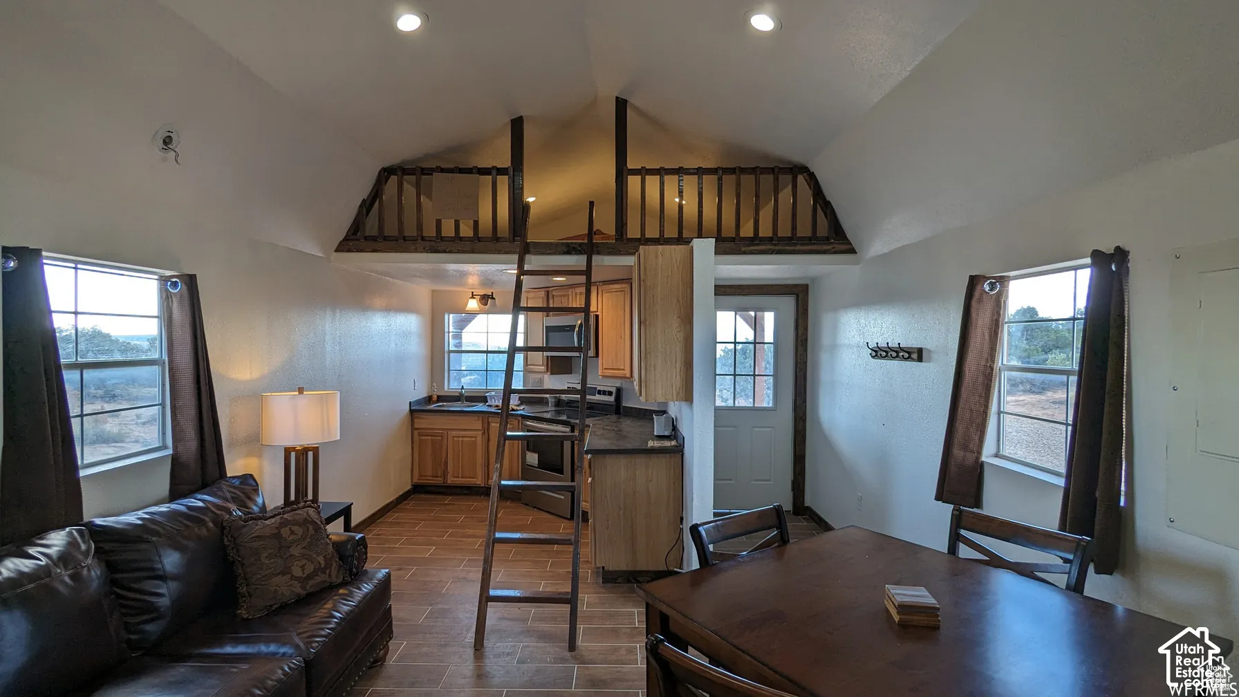 Vaulted ceilings with recessed lighting, an elevated sleeping loft featuring a wood railing, tiled flooring, wood cabinetry, and multiple windows providing natural light
