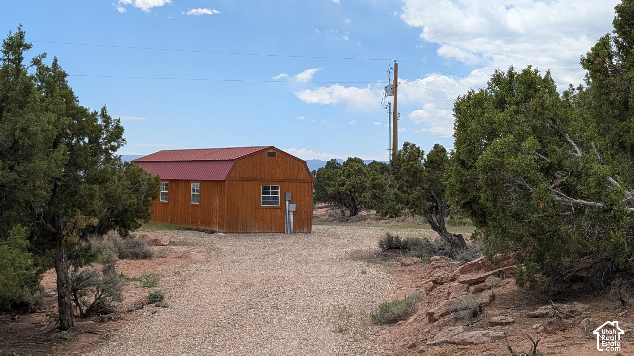 Wood-sided structure featuring a metal roof, slider windows, and a gravel path, situated on a lot with native trees and rock outcroppings
