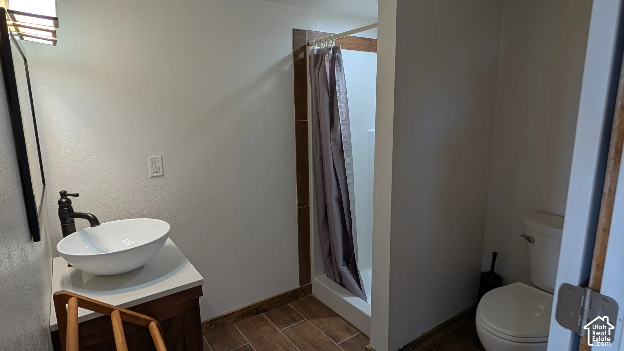Bathroom featuring a vessel sink with a dark-finish faucet, white countertop, wood-finish vanity cabinetry, wood-finish tile flooring, and a tiled shower surround