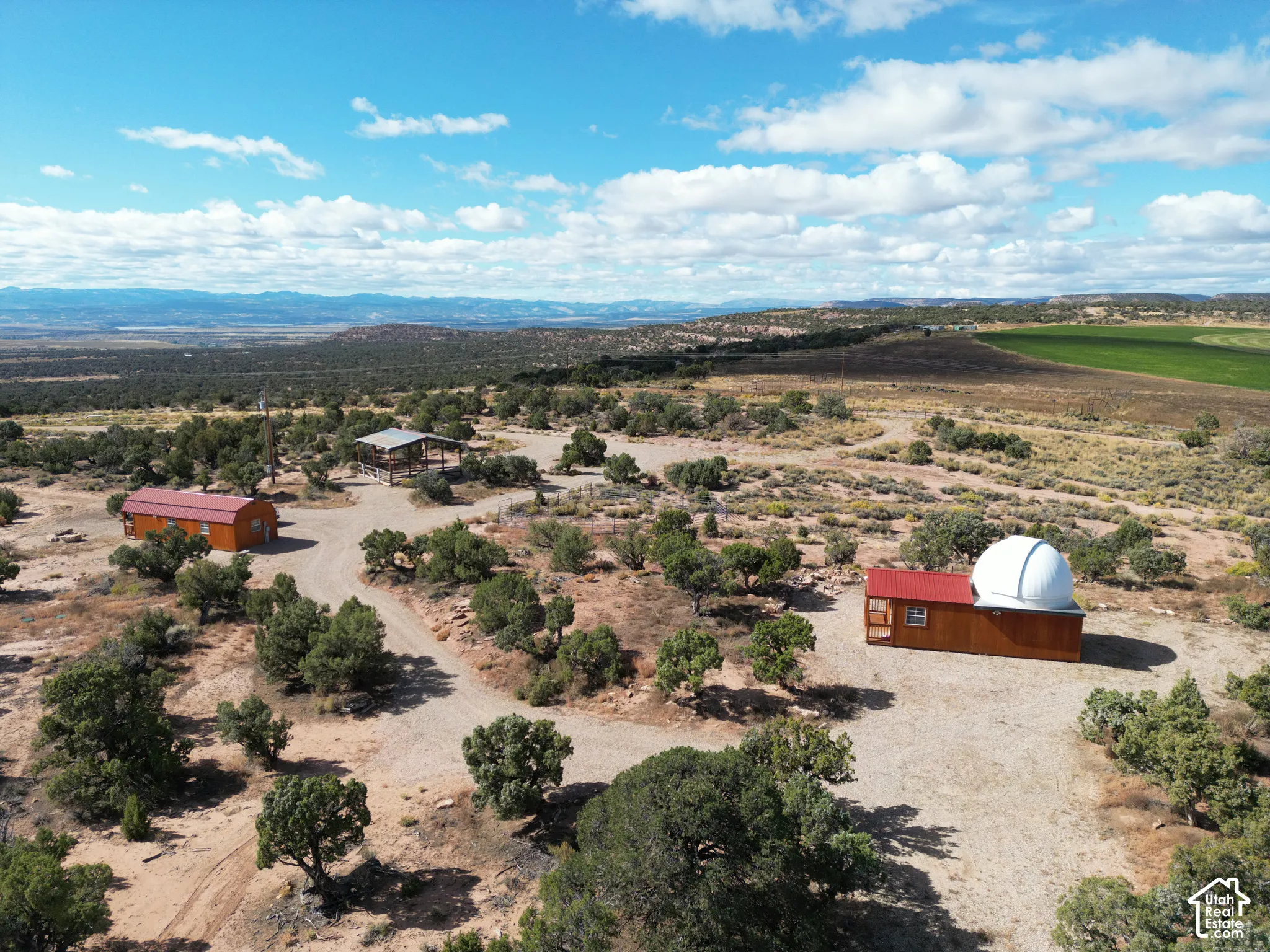 Aerial perspective of a sprawling property featuring multiple structures, including a wood-sided building with an integrated observatory dome, a detached rectangular outbuilding with a metal roof, an open-sided pavilion, and expansive native desert landsc