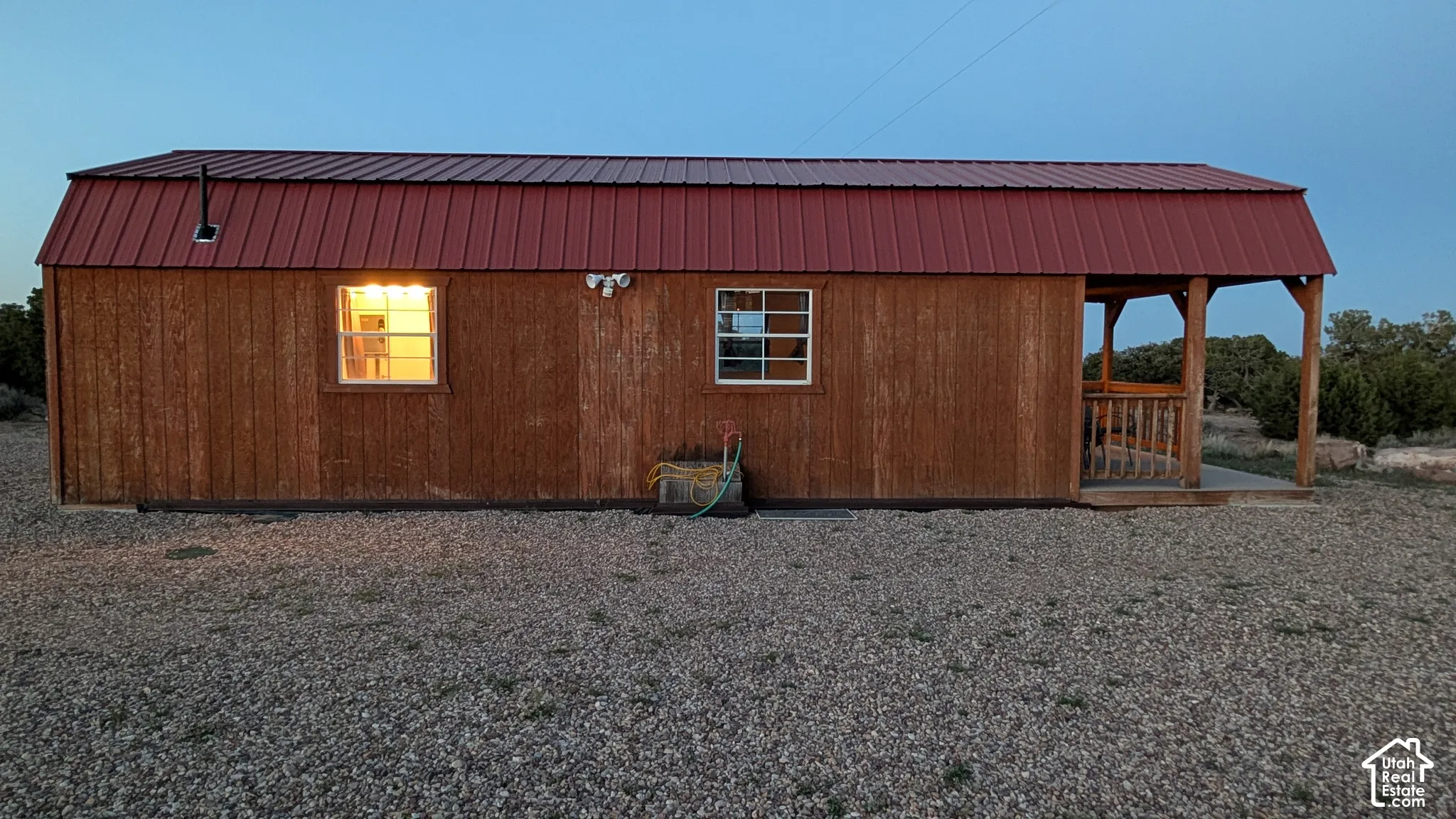 Single-story structure featuring wood-finish siding, a gambrel roofline with red metal roofing, a covered porch with wooden railings, and grid-pane windows
