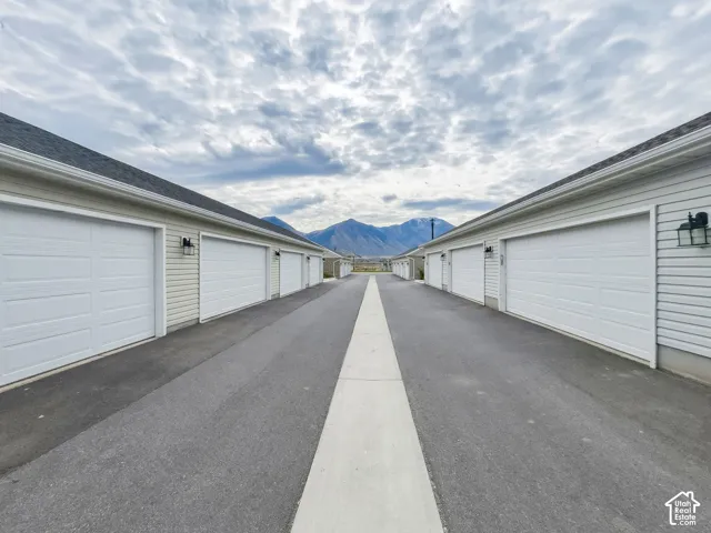 Paved access alley lined with multiple white garage doors, siding exterior, attached carriage-style lighting, and a mountain backdrop
