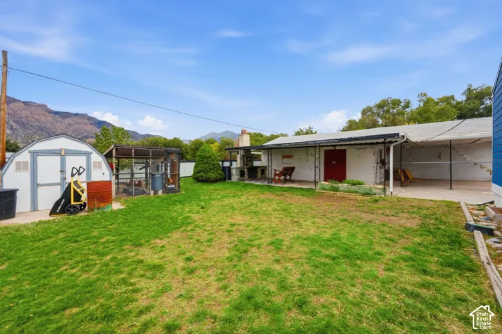 Backyard featuring a grass lawn, paved patio area, covered outdoor structure, masonry chimney, and storage shed