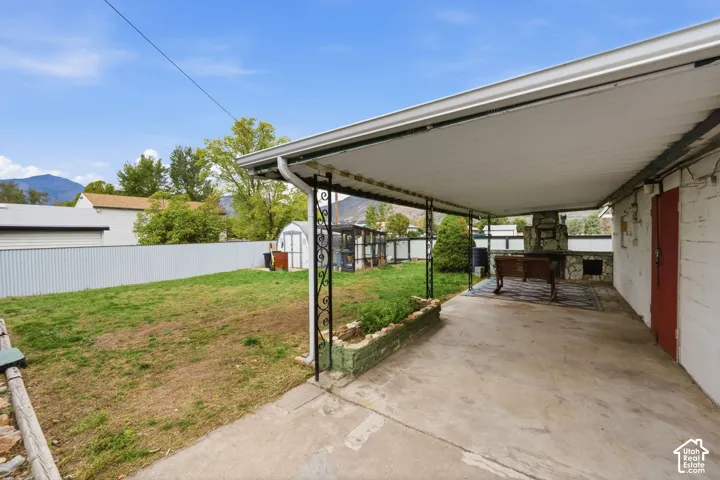 Covered concrete patio with decorative metal supports, stone-faced masonry fireplace, raised brick garden bed, and enclosed backyard featuring metal perimeter fencing