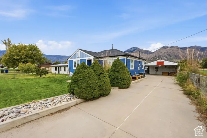 Exterior featuring blue lap siding, an expansive concrete driveway, a detached carport, a landscaped front lawn with stone-edged garden beds, and a mountain backdrop