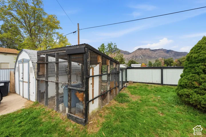Enclosed poultry structure with black-finished framing, metal mesh siding, and shingled roof, set on a grass-covered lot with perimeter mesh fencing and mountain backdrop