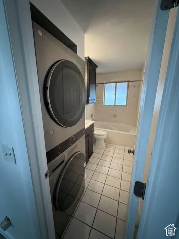 Stacked front-load laundry appliances, dark wood-finish cabinetry, tile flooring, and a shower-tub combination with a window