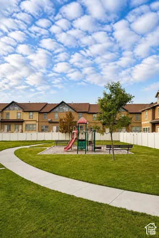 Communal greenspace featuring a concrete pathway, manicured lawn, playground equipment, and perimeter vinyl fencing