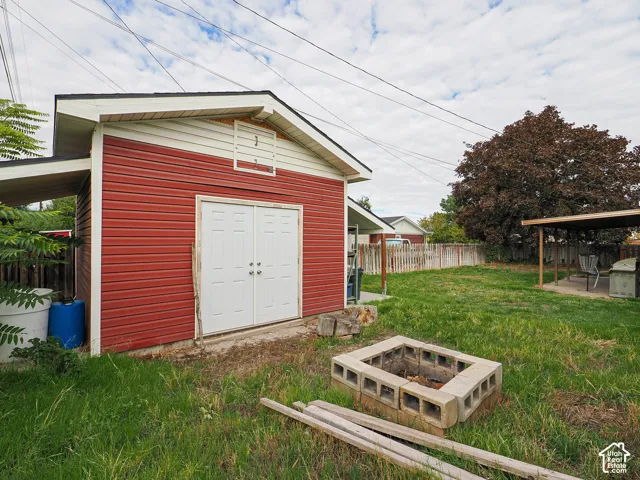 Detached storage structure featuring horizontal red siding, gable roof, and double entry doors, accompanied by a concrete block fire pit and a covered patio area set within a fenced yard