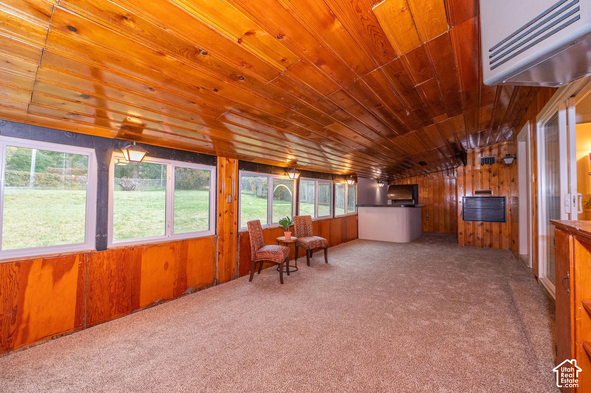 Enclosed sunroom featuring natural wood-paneled walls and ceiling, carpeted flooring, large multi-pane windows, and a built-in wet bar with a countertop