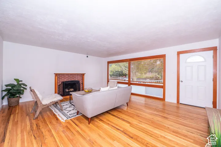 Living area featuring hardwood flooring, brick fireplace with a wood mantel, large picture windows with wood trim, and a white entry door