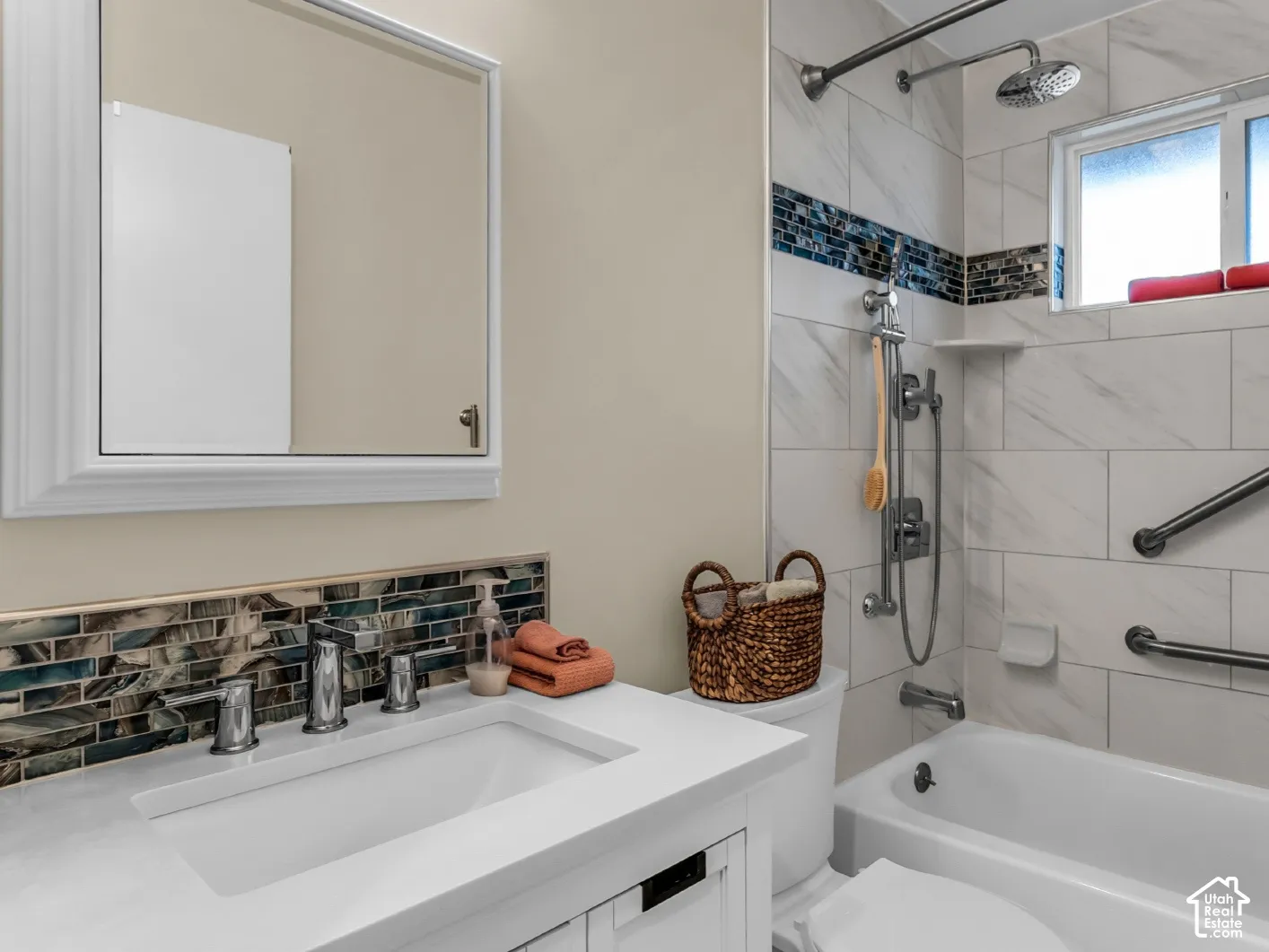 Bathroom featuring a white vanity with a solid-surface countertop, rectangular basin, mosaic tile backsplash, framed wall mirror, and a tiled tub-shower combination with integrated wall-mounted grab bars and a handheld showerhead