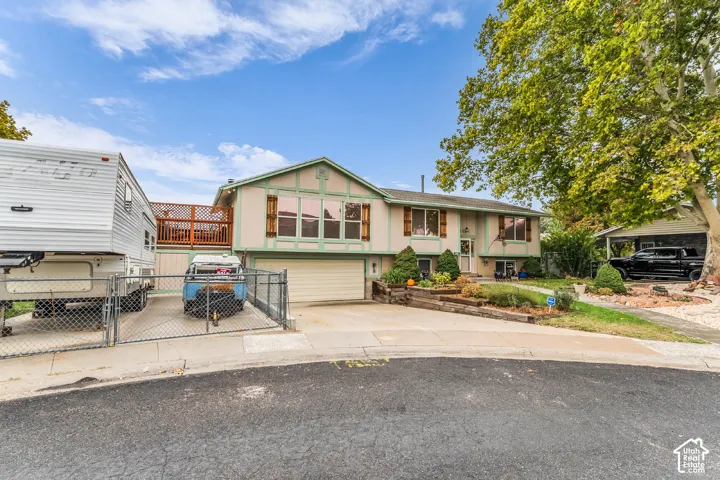 Split-level exterior featuring a paved driveway, attached garage, elevated wood deck with lattice railing, and tiered timber landscape retaining walls
