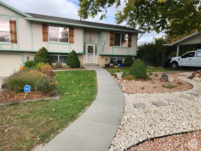 Split-level exterior featuring wood-accented shutters, concrete walkway, decorative stone landscaping, attached carport, and a manicured lawn
