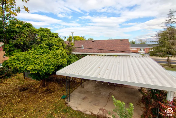 Covered metal-roofed carport with concrete flooring, asphalt shingle roofing, and mature shade trees