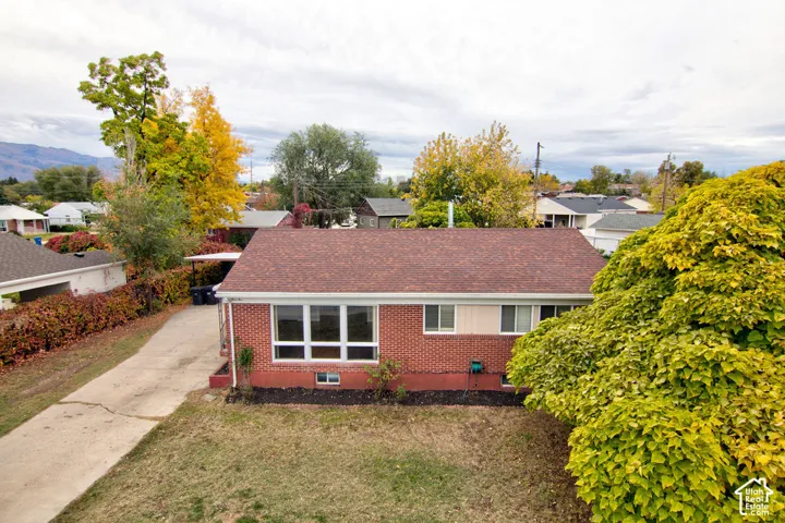 Single-story exterior featuring a brick facade, shingle roof, large multi-pane front window, concrete driveway, and surrounding mature trees