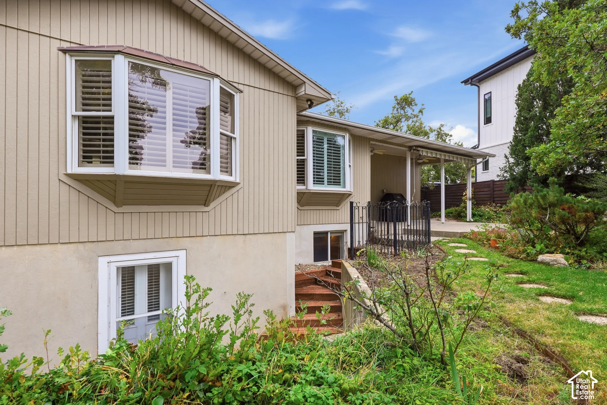 Vertical siding exterior featuring two bay windows, a covered patio with metal railing, and stone-paver walkway.