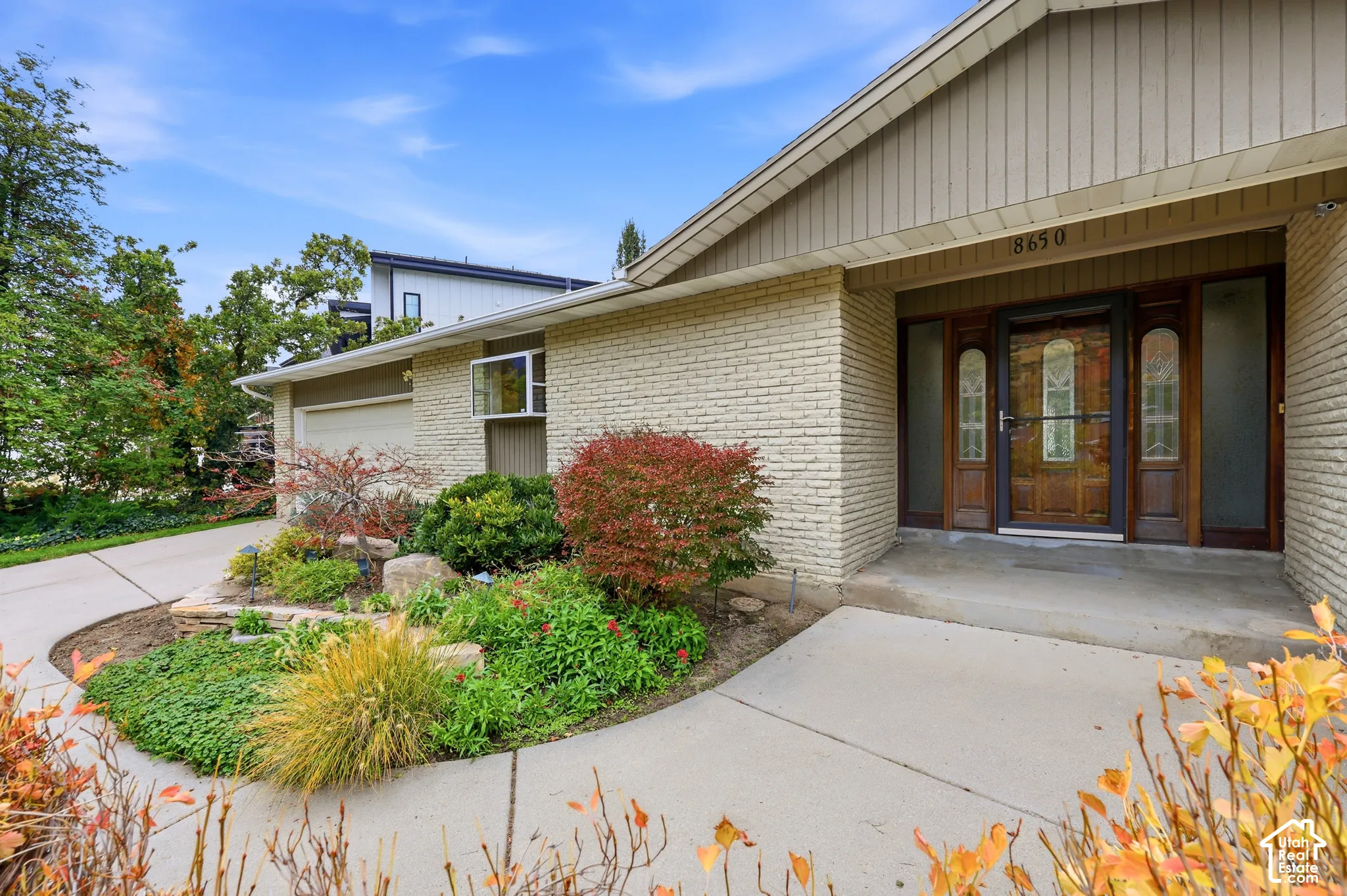 Cream-colored brick exterior featuring a recessed entryway, ornate wood-frame front door with glass sidelights, and a concrete driveway leading to a multi-car garage. Color correction applied.