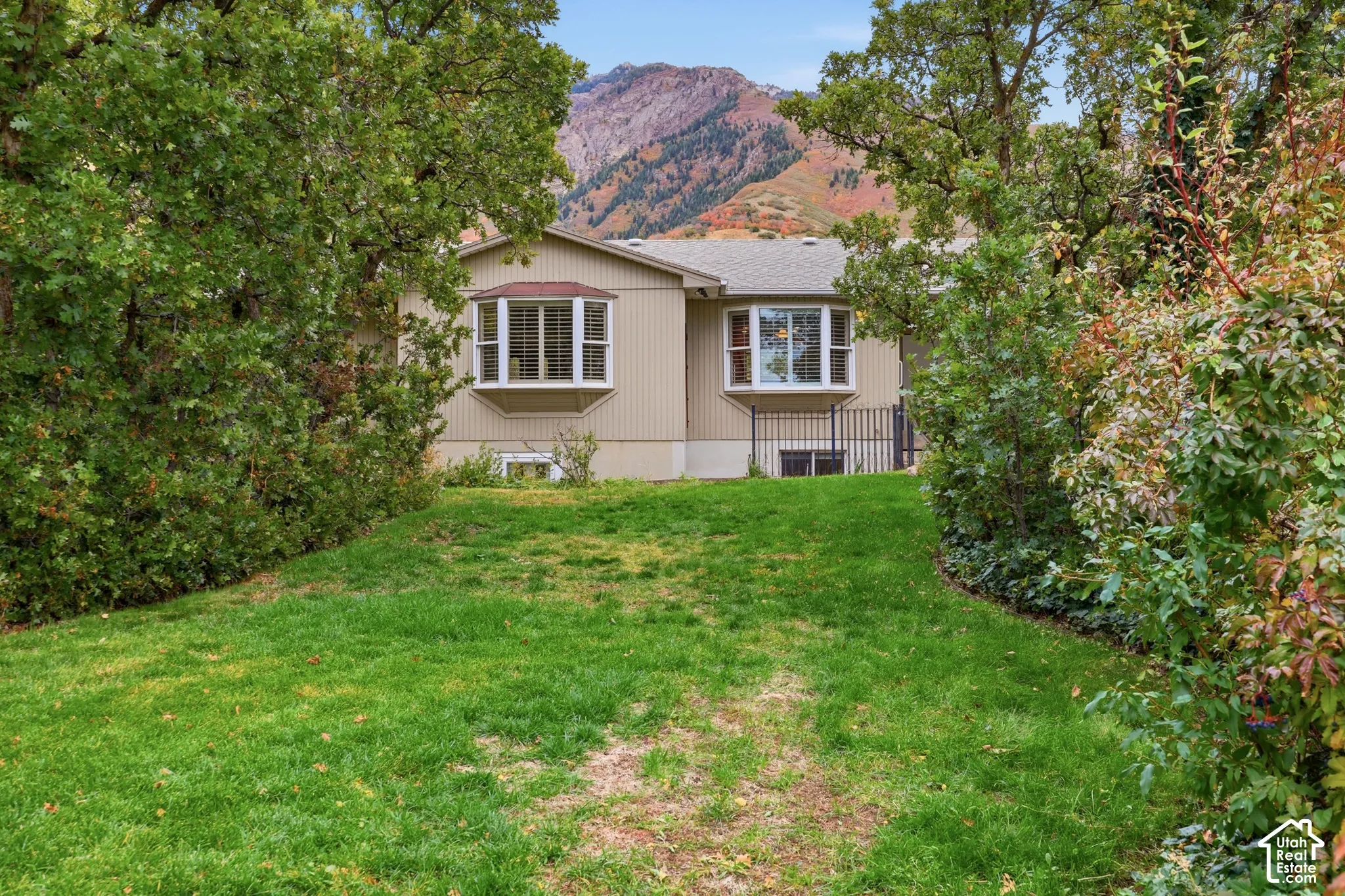 Exterior featuring vertical siding, dual bay windows, mature tree-lined landscaping, and a mountain backdrop.