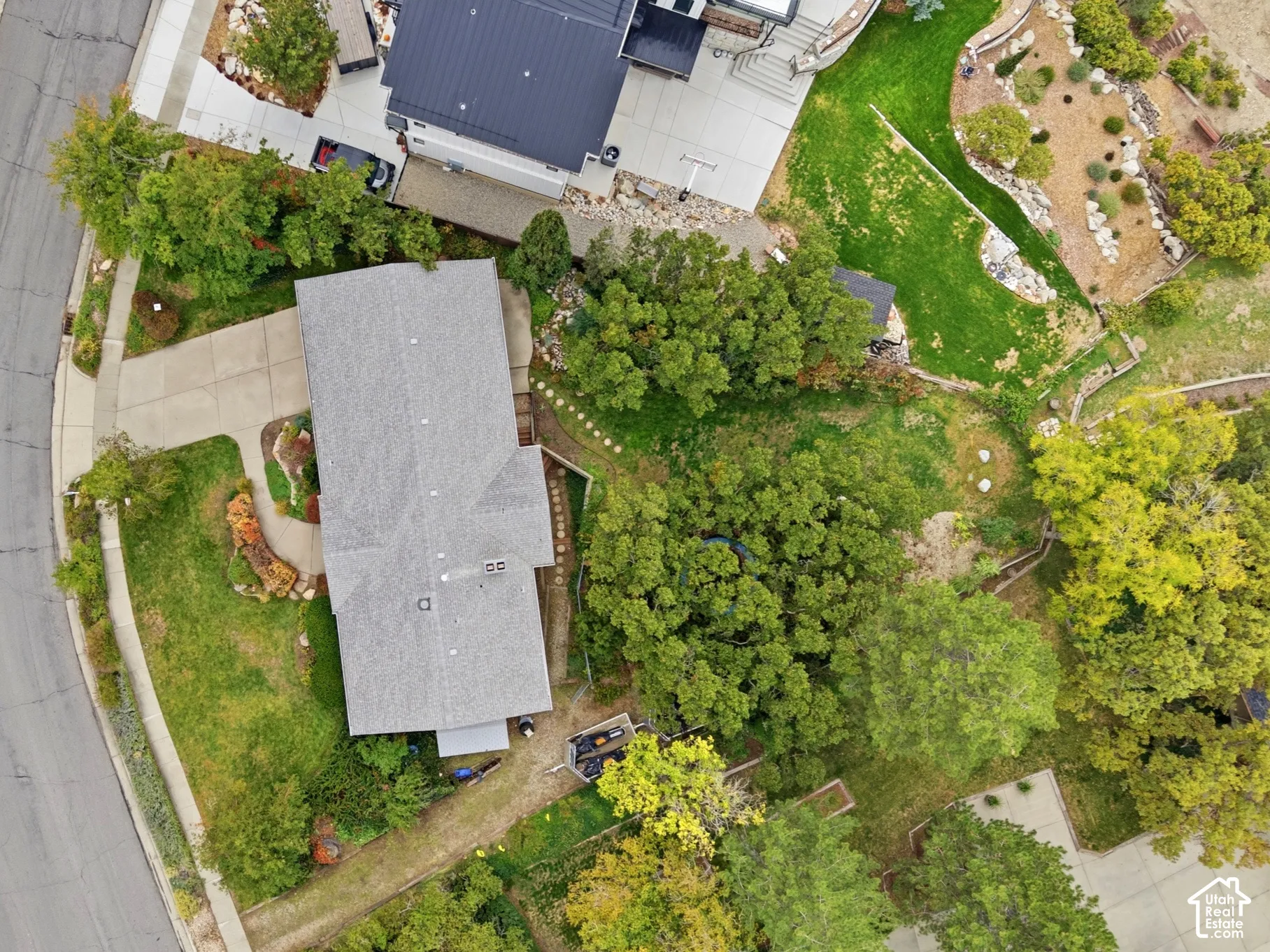 Aerial perspective featuring a pie shaped lot with a concrete driveway, multi-level landscaping, mature trees, and asphalt shingle roofing.