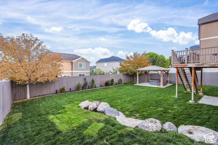 Fenced backyard featuring a manicured lawn, stone hardscaping, raised wood deck with staircase, and a covered pavilion