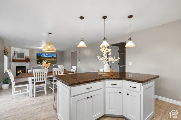 Kitchen island with stone-surface countertop, white shaker-style cabinetry, triple pendant lighting, wood-finish flooring, and wall-integrated fireplace with a wood mantel