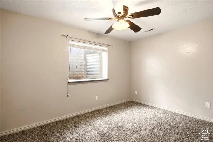 Neutral-toned interior featuring textured walls, wall-to-wall carpeting, a multi-blade ceiling fan with integrated lighting, and a window with a window well