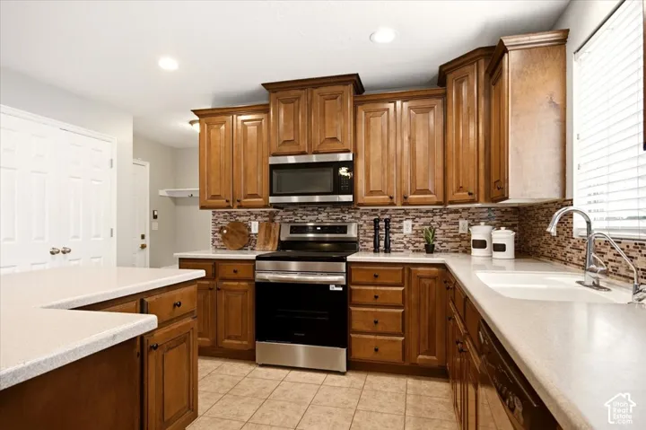 Kitchen featuring medium-tone wood cabinetry, solid-surface countertops, a mosaic tile backsplash, stainless steel appliances, and tile flooring