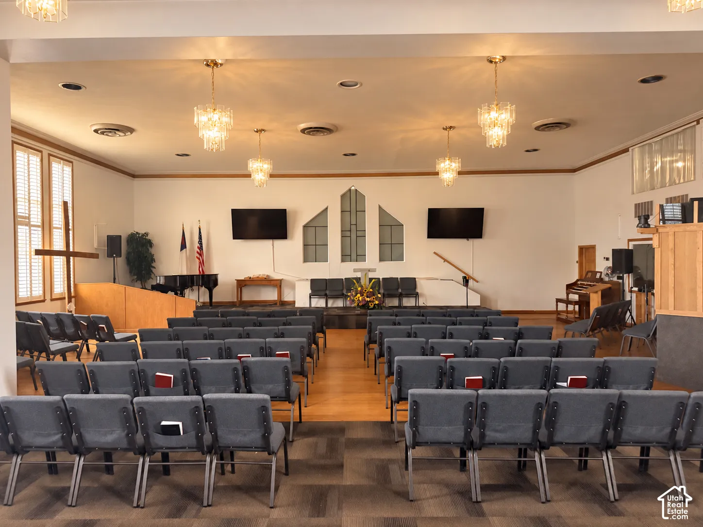 Large assembly hall featuring a raised stage, multiple crystal-style chandelier fixtures, decorative window inserts, wood-finish flooring, and wall-to-wall carpeting