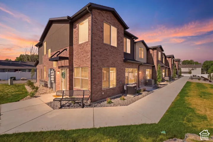 Multi-story brick exterior featuring dark trim, large windows, concrete walkways, and established landscape beds