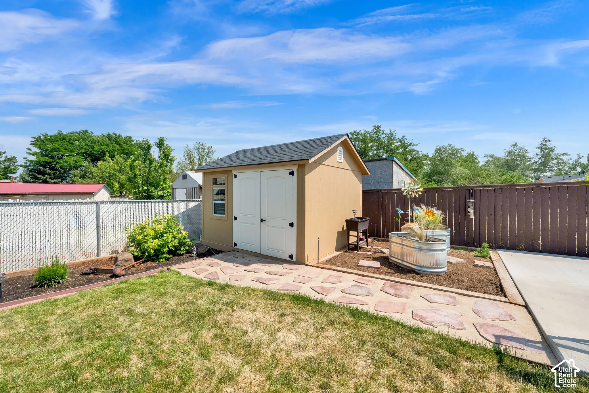 Detached storage shed with dual doors and shingled roof, accented by a flagstone walkway, concrete patio, and perimeter wood fencing
