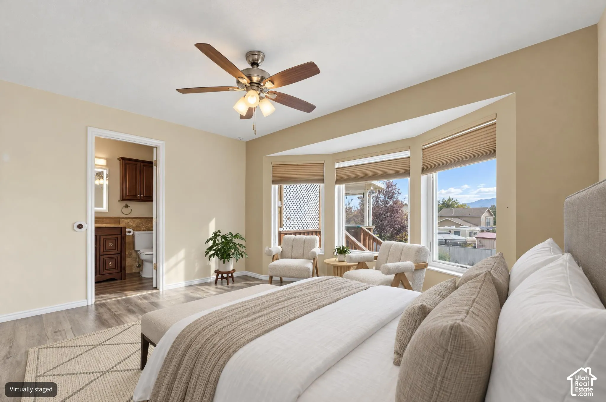 Digitally altered bedroom featuring a bay window, wood-finish flooring, and a multi-blade ceiling fan with integrated lighting