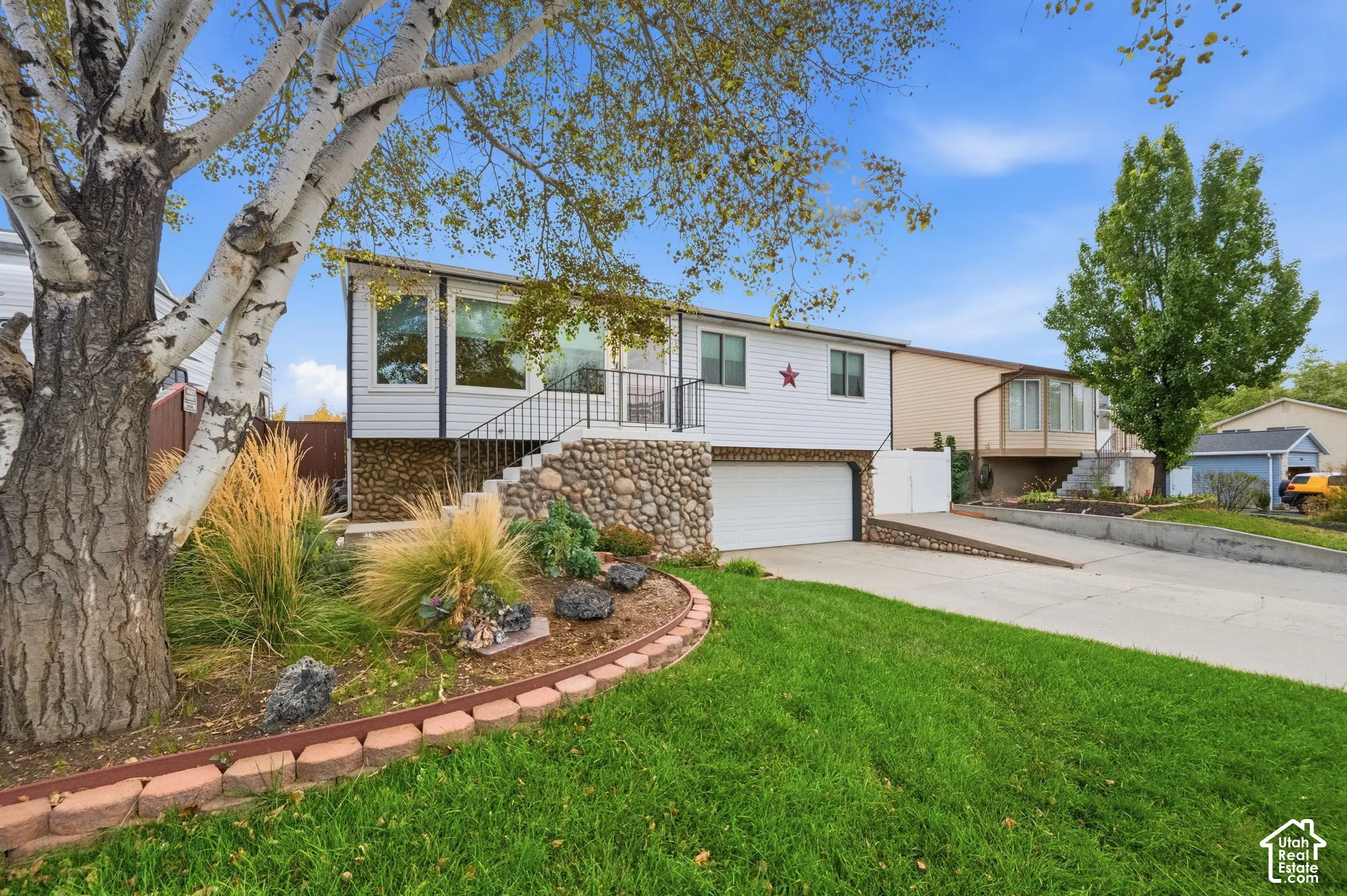 Exterior facade featuring white horizontal siding, stone-veneer foundation, attached double-car garage, concrete driveway, and landscaped front yard with stone edging