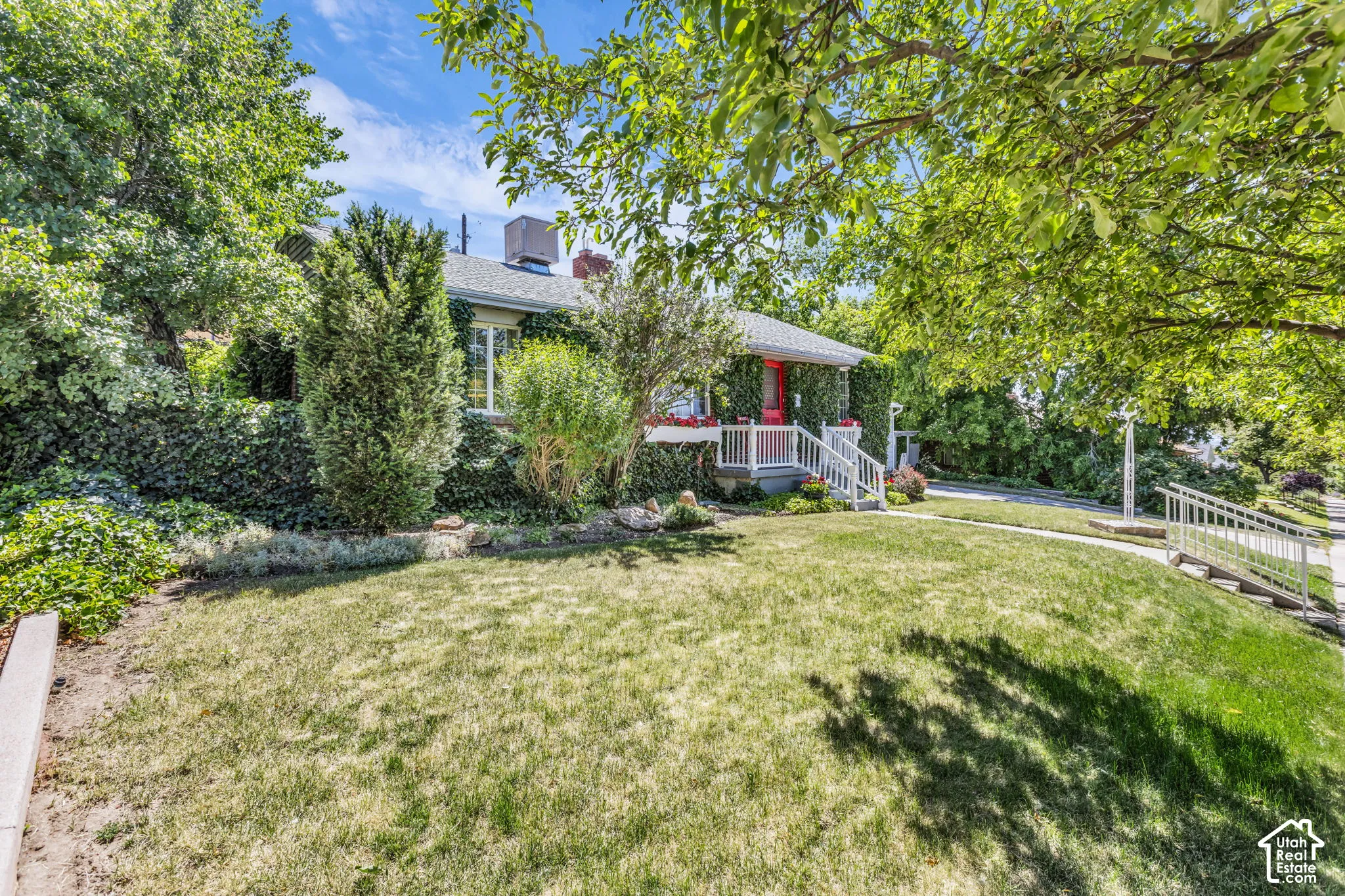Single-story exterior featuring a brick facade, white railing along the front porch, sloped roofline, mature trees, and tiered landscaping