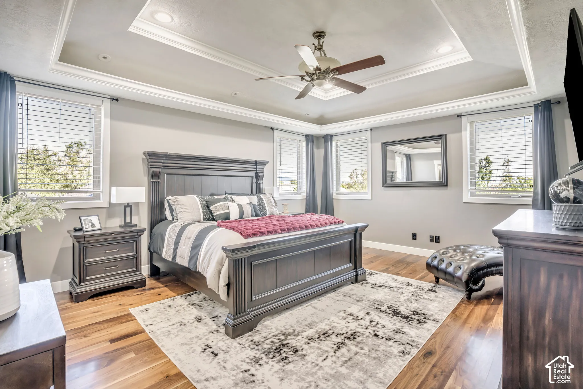 Tray ceiling with recessed lighting, ceiling fan, wood-finish flooring, and multiple windows with horizontal blinds