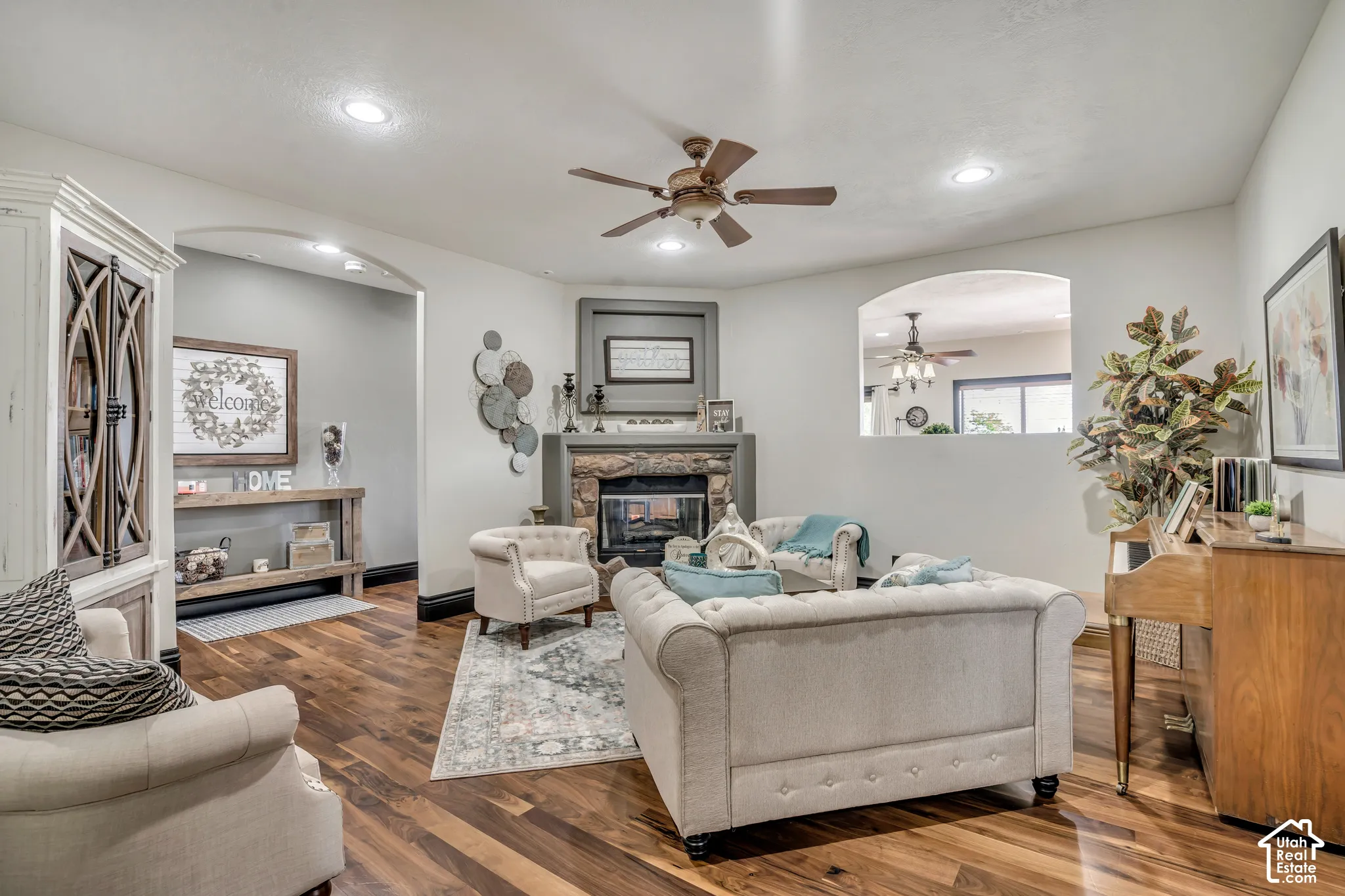 Living area featuring wood-finish flooring, stone-surround corner fireplace, recessed lighting, ceiling fan, and arched architectural openings