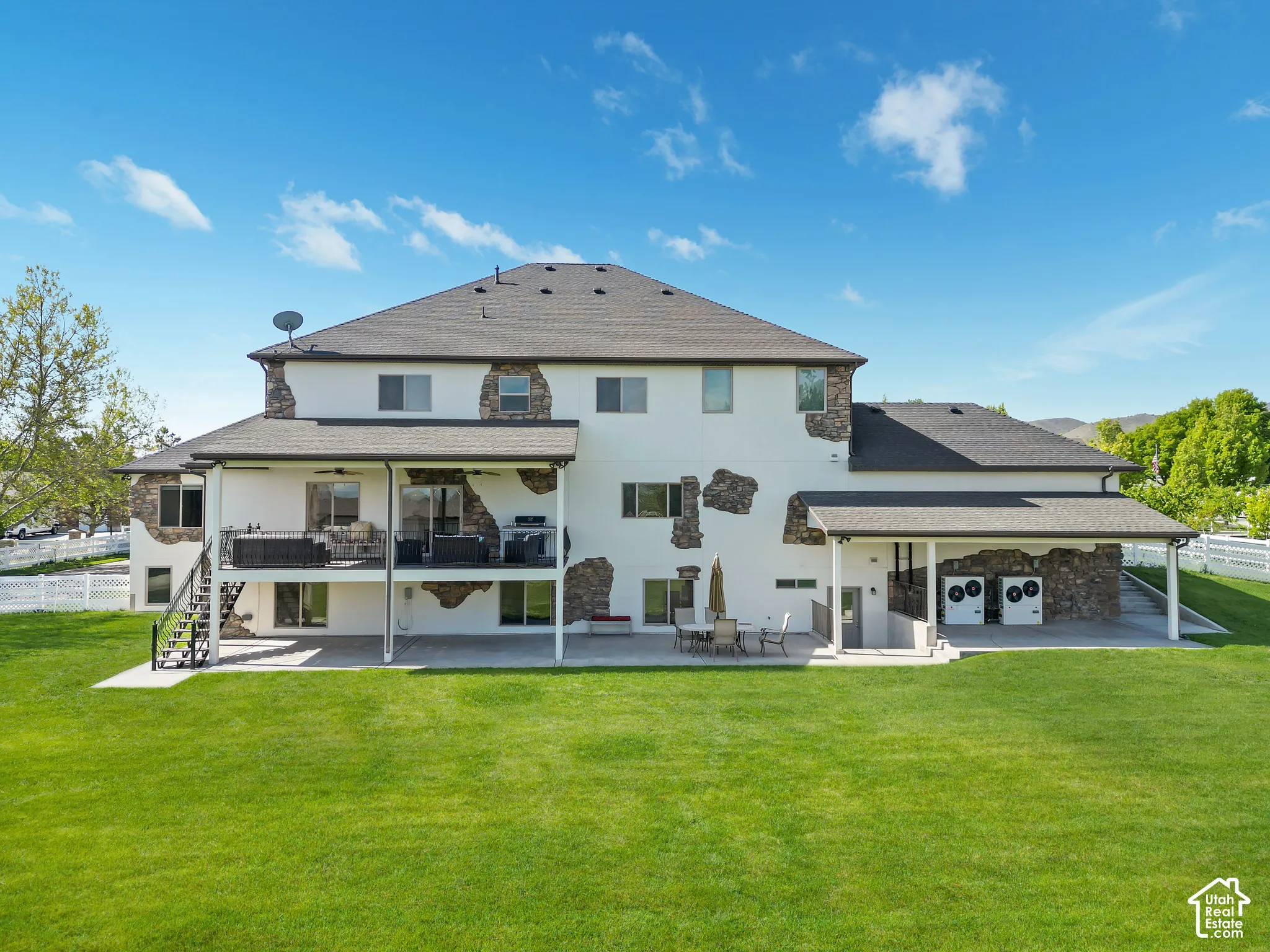 Rear exterior featuring a stone and stucco facade, a covered elevated deck with metal railing, a ground-level concrete patio, and a grass lawn enclosed by white vinyl fencing
