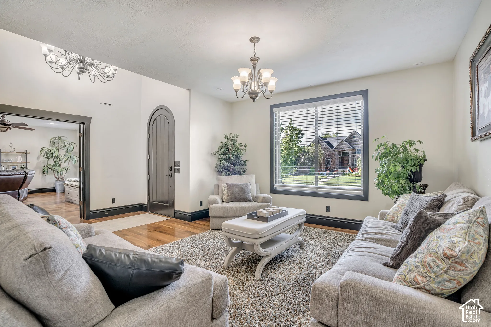 Arched wooden front entry door, wood-finish flooring, high ceilings with dual classic-style chandeliers, and dark-finished window and door casings