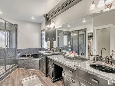 Dual-sink vanity with stone countertops, corner soaking tub with stone surround, walk-in glass-enclosed shower, expansive wall mirror, and wood-finish flooring